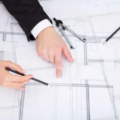 Dwie osoby analizują projekt budowlany, dyskutując, jak zaznaczyć zmiany nieistotne. Na stole leżą cyrkiel, ołówki i linijka.