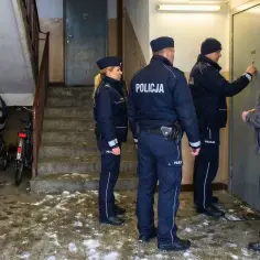 Policja puka do drzwi, by sprawdzić, czy nadzór budowlany może wejść do mieszkania. Na klatce schodowej stoją wózki i rowery.
