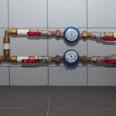 Dwa wodomierze radiowe w rzędzie, z czerwonymi zaworami. Instalacja wodna z mosiężnymi złączkami i białymi rurami.