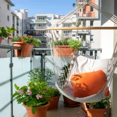 Przytulny mini balkon w bloku z wiszącym fotelem, poduszkami i kwitnącymi roślinami. Idealne miejsce na relaks.