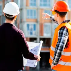 Dwóch budowlańców w kaskach analizuje plany. Jeden z nich trzyma zwinięty projekt, drugi wskazuje kierunek. Czy to zagubiony dziennik budowy?