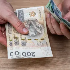 Dłonie liczą polskie banknoty. Czy mieszkaniówka wlicza się do dochodu? Pieniądze na drewnianym blacie.