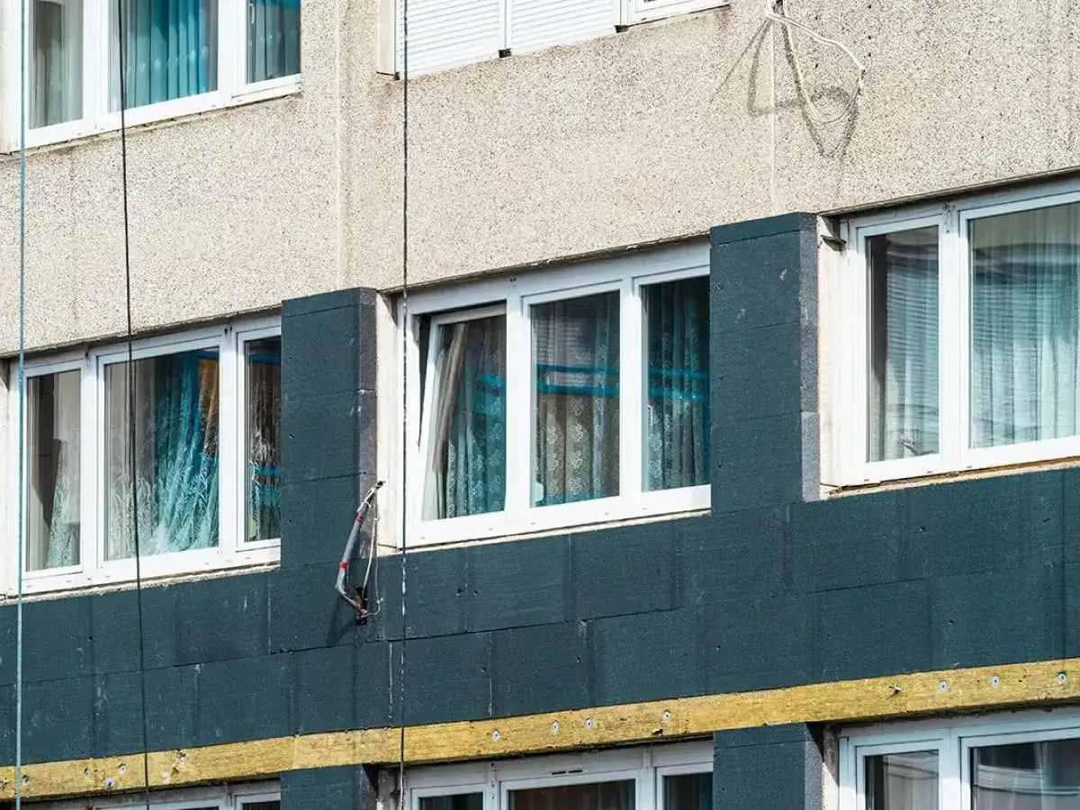 Termomodernizacja mieszkania w bloku: ciemna izolacja na ścianie, okna z zasłonami, żółty pas izolacji.