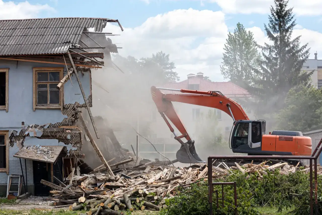 Koparka pracuje przy rozbiórce domu, unosząc tumany pyłu. To dobry przykład, jak zrobić opis rozbiórki.