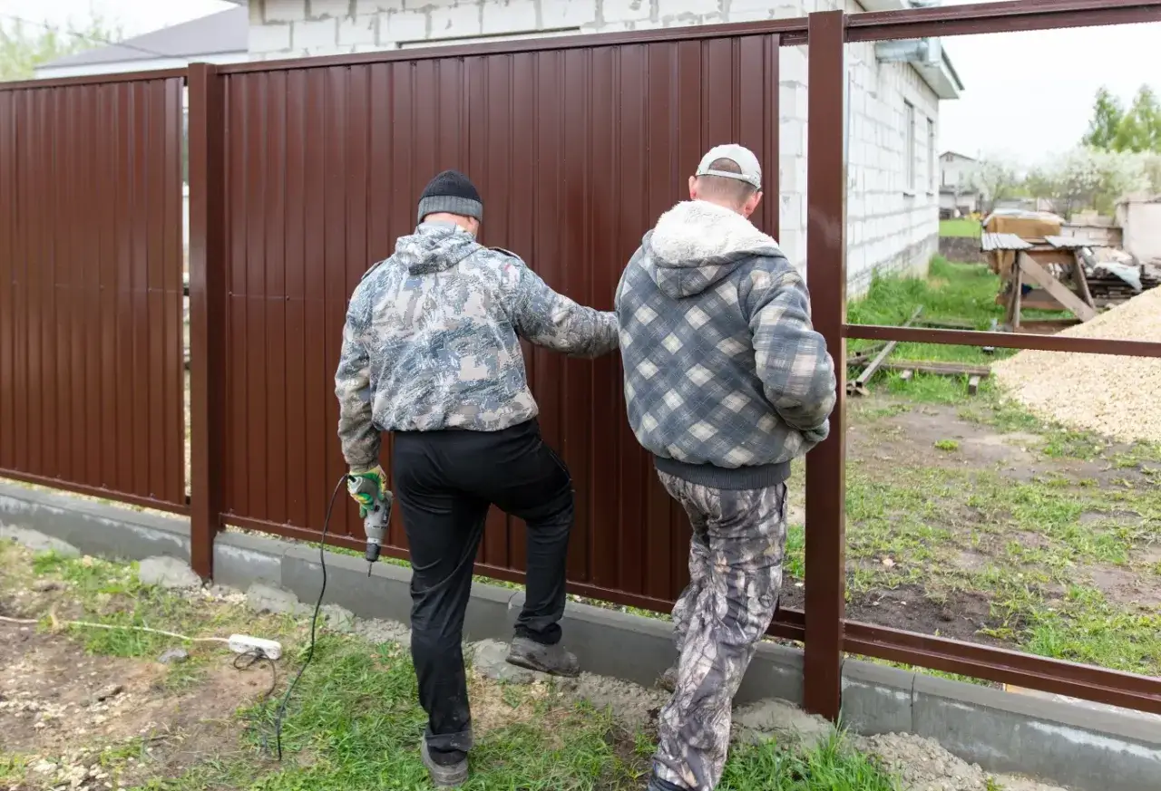 Dwóch mężczyzn montuje brązowe ogrodzenie. Jeden trzyma wiertarkę. To część prac budowlanych, o których sąsiedzi zostali poinformowani.